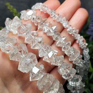 Peut inclure: Gros plan sur des perles de quartz transparentes et facettées. Les perles, de forme irrégulière, sont enfilées pour former un collier ou un bracelet. Le quartz reflète la lumière. L'arrière-plan est flou, avec des nuances de vert et de violet.