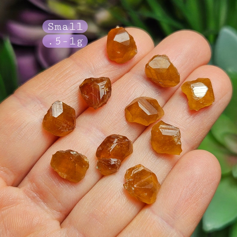 May include: A collection of small, rough-cut, orange-brown gemstones, each approximately .5-1g in weight, are displayed in a hand. The gemstones have a faceted appearance and varying shapes. The word "Small" is written above the weight range.
