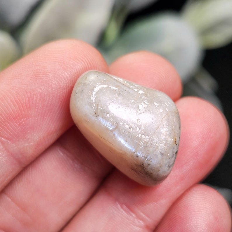 Puede incluir: Una gema pulida, de forma ovalada, con un brillo nacarado, sostenida en una mano. La piedra es de color gris claro con sutiles variaciones y una superficie lisa y redondeada. Es un objeto natural.