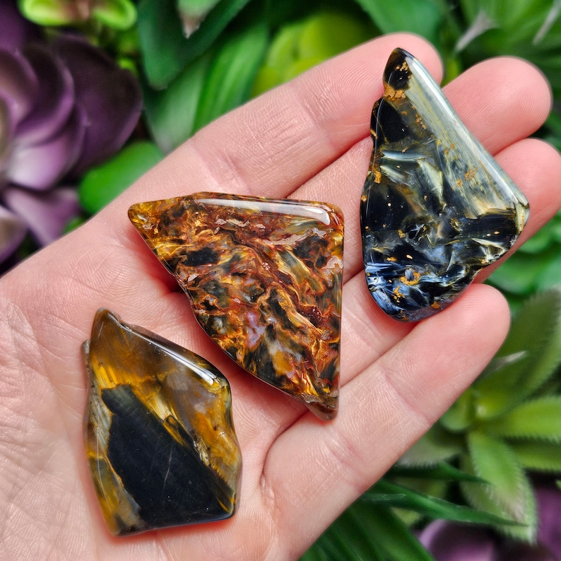May include: Three polished, irregularly shaped gemstones with smooth, reflective surfaces. One stone features deep black and golden-brown bands, another displays a swirling mix of amber, gold, and black, and the third shows blue, black, and gold.