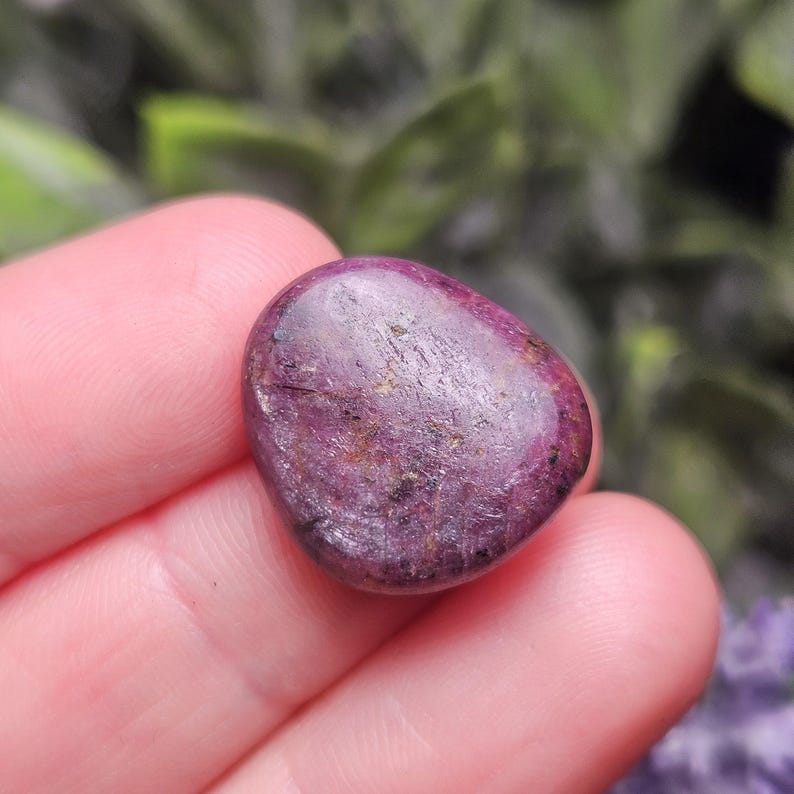 May include: A polished, oval-shaped gemstone in shades of deep red and purple, held in a hand. The stone has a smooth, reflective surface with subtle variations in color and texture. The background is blurred, with hints of green and purple.