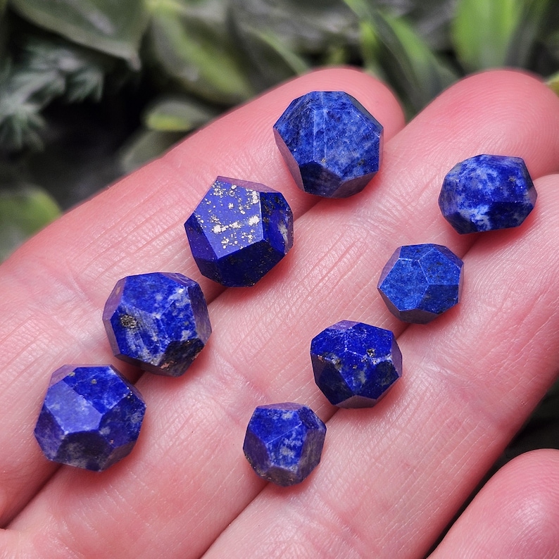 May include: A collection of seven small, faceted lapis lazuli gemstones. Each stone displays a deep blue color with natural white and gold-colored mineral inclusions, cut into irregular geometric shapes with multiple flat, polished surfaces.