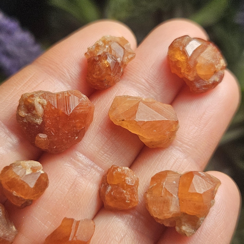 May include: A collection of raw, orange-brown garnet crystals. The gemstones have a rough, natural texture and exhibit various faceted shapes. The crystals are displayed against a blurred background, highlighting their color and form. These gemstones are ideal for jewelry making or collecting.
