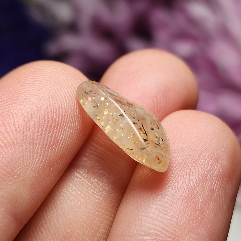 May include: A polished, teardrop-shaped gemstone with a translucent, golden-yellow hue. The stone is speckled with shimmering gold and dark inclusions, creating a unique pattern. The gemstone is held in a hand.