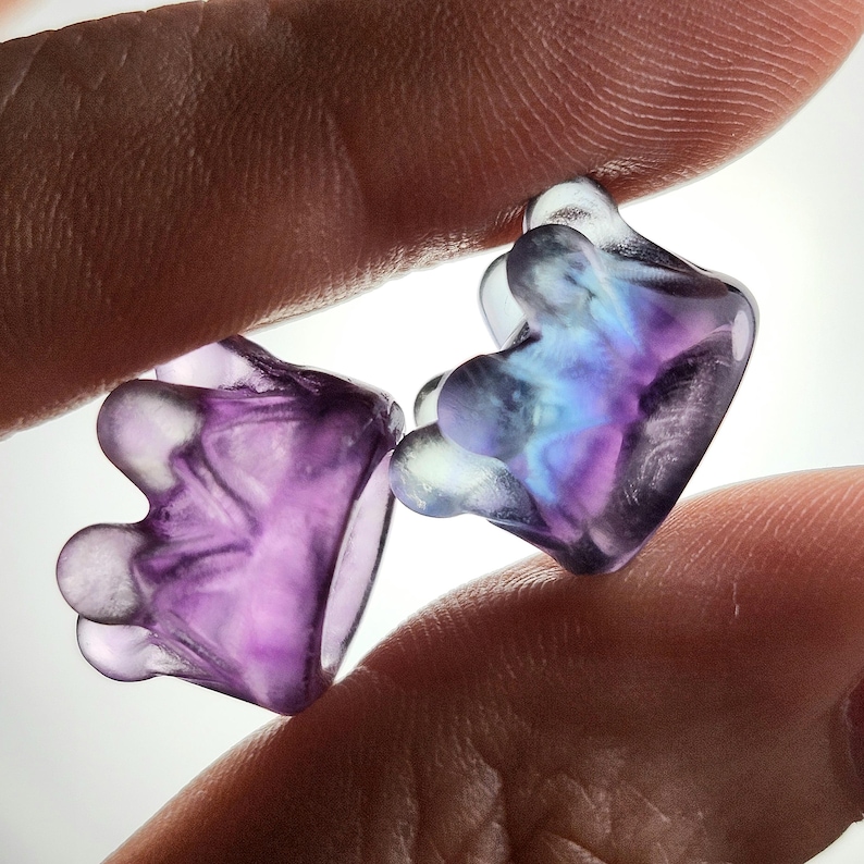 May include: Two translucent, abstract-shaped earplugs with a unique design. The earplugs are held between fingers and display a blend of purple, blue, and clear colors. The overall shape is reminiscent of a stylized paw print.