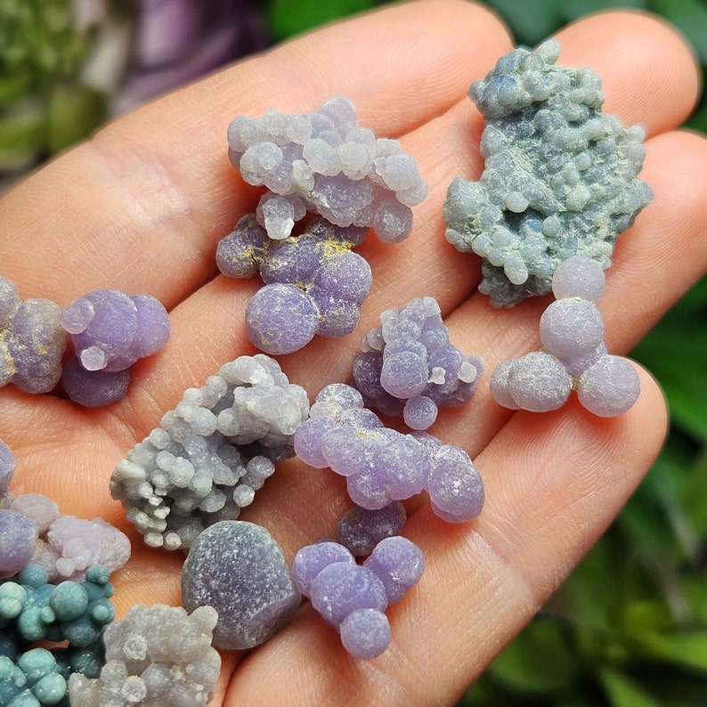 May include: A selection of small, grape-like clusters of polished gemstones in shades of lavender, purple, and grey. The stones are held in a hand, with a blurred green background. The gemstones have a smooth, rounded texture and vary in size and shape.
