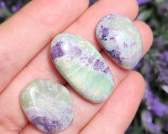 Pode incluir: São exibidas três pedras preciosas polidas em tons de verde e roxo. As pedras têm formato oval ou arredondado. As pedras preciosas são seguradas em uma mão, com um fundo desfocado de flores roxas.