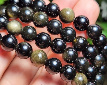 May include: A close-up of a black and gold beaded bracelet. The beads are round and have a shiny finish. The bracelet is made of multiple strands of beads.