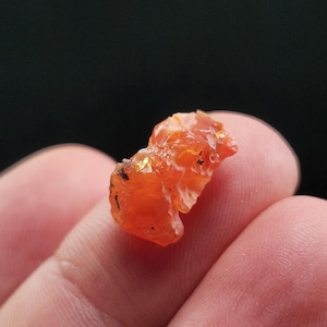 May include: A rough, orange-colored gemstone with a crystalline structure. The gemstone has a translucent appearance and is held between fingers against a dark background. The stone has a rough, uneven surface.