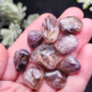 May include: A collection of polished amethyst stones in various shades of purple, brown, and white. The stones are smooth and oval-shaped, with natural patterns and textures. The gemstones are held in a hand, showcasing their size and color variations.