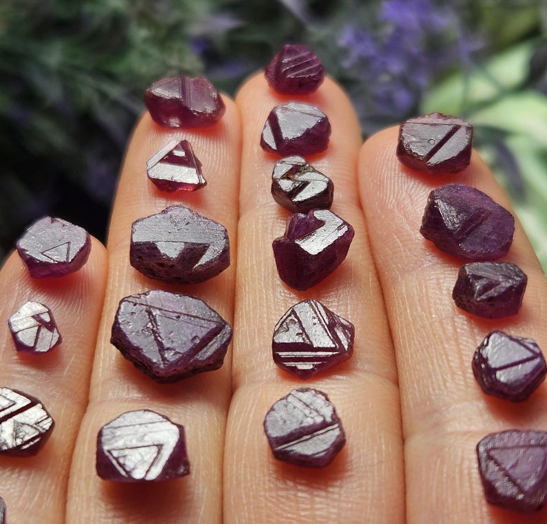 K&ouml;nnte beinhalten: Eine Sammlung von 16 kleinen, dunkelvioletten, facettierten Rubinkristallen. Die Kristalle sind auf einer Hand angeordnet, wobei sich einige Kristalle &uuml;berlappen.