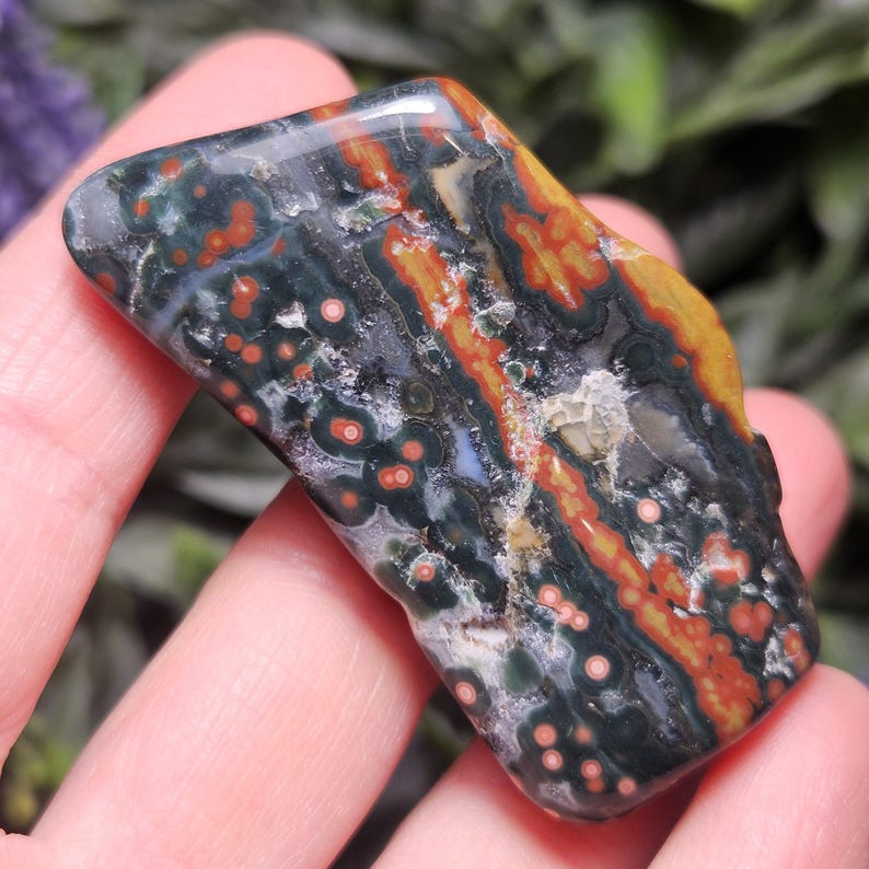 Puede incluir: Una piedra de jaspe oce&aacute;nico pulida con una base de color verde oscuro y patrones orbiculares distintivos en tonos rojos, naranjas y amarillos. La superficie muestra una textura suave y brillante con inclusiones minerales naturales.