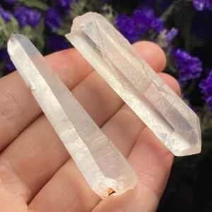 May include: Two clear quartz crystal points, one slightly larger than the other, with a natural, raw finish. The crystals are held in a hand, showcasing their length and shape.