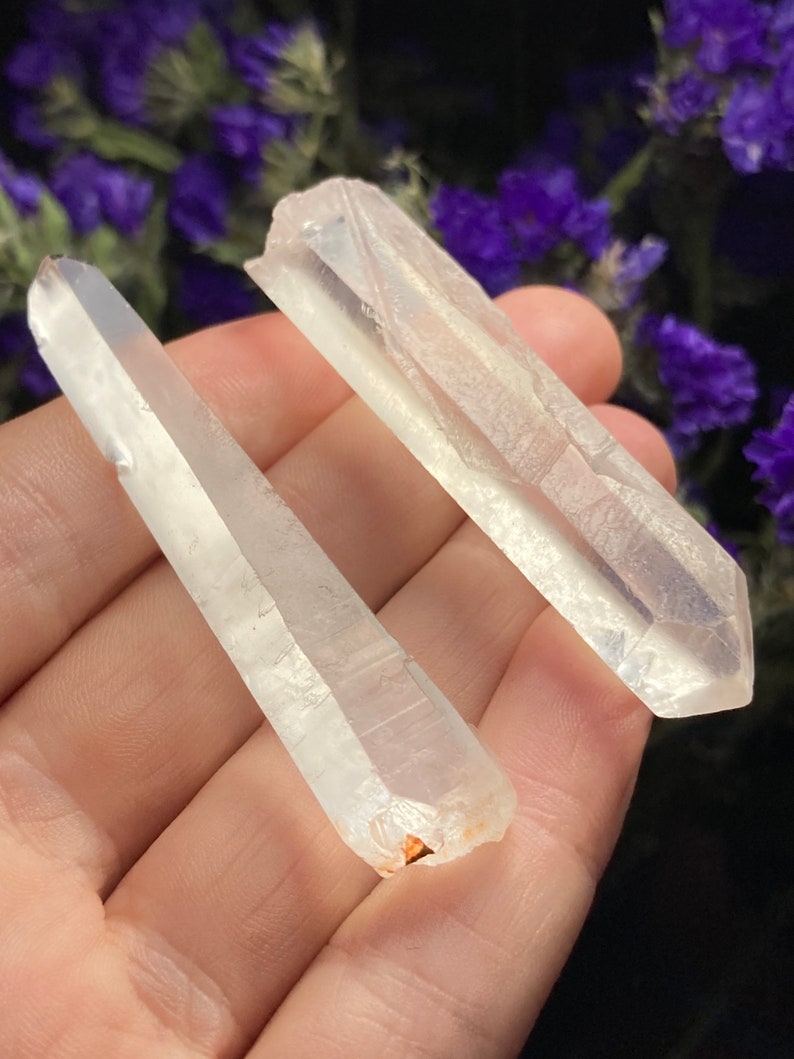 May include: Two clear quartz crystal points, one slightly larger than the other, with a natural, raw finish. The crystals are held in a hand, showcasing their length and shape.