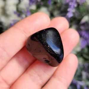 May include: A polished, triangular-shaped black stone with a smooth, reflective surface and several small, natural indentations or pits on its front face. The stone has a deep, solid black color with subtle light reflections on its surface.