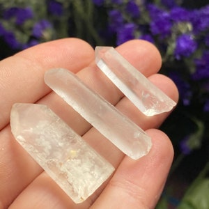 May include: Three clear quartz crystal points, also known as rock crystals, are held in a hand. The crystals are translucent and have a natural, raw appearance.