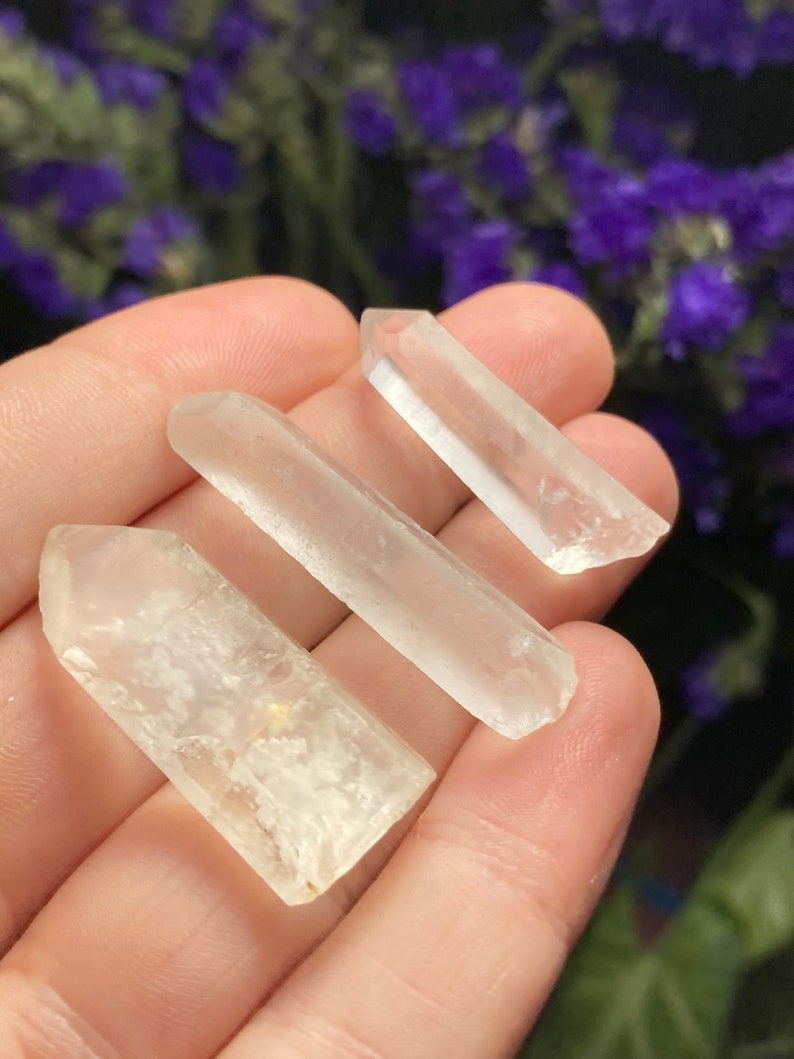 May include: Three clear quartz crystal points, also known as rock crystals, are held in a hand. The crystals are translucent and have a natural, raw appearance.
