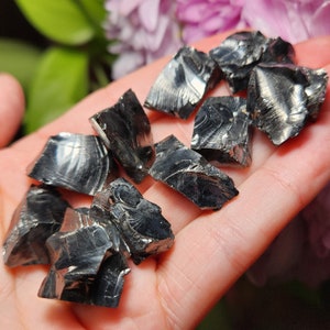 May include: A collection of black obsidian crystals, also known as volcanic glass, with a shiny, iridescent surface. The crystals are various shapes and sizes, and are displayed on a hand.