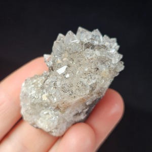 May include: A cluster of clear quartz crystals with sharp, pointed terminations. The crystals are a translucent, off-white color with some inclusions. The quartz is held in a hand against a black background.