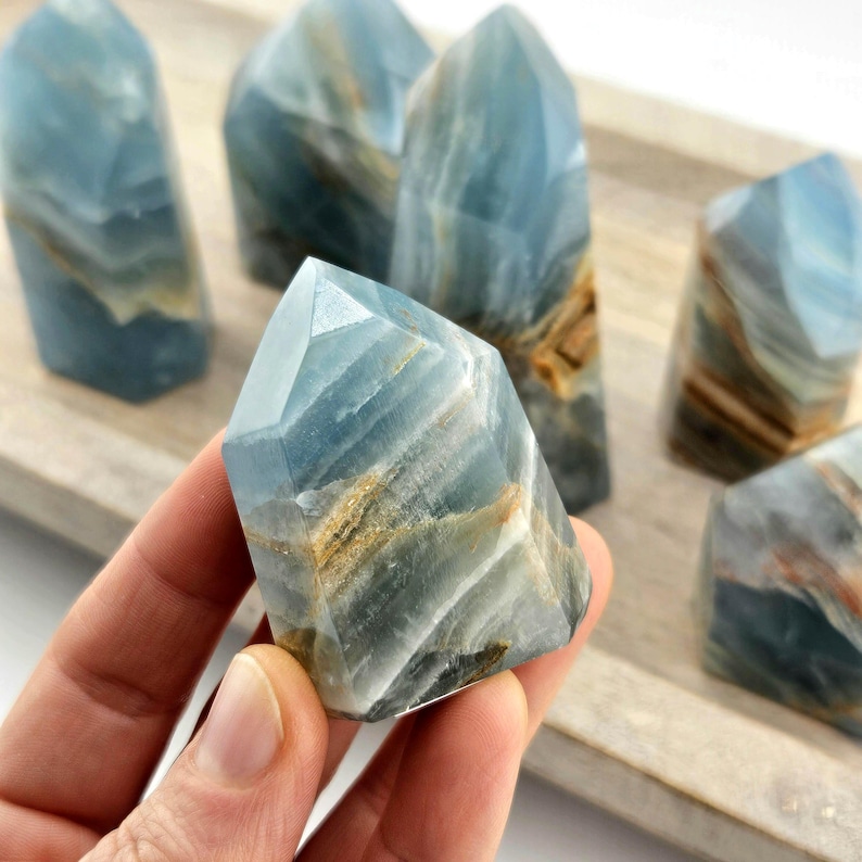 May include: A selection of blue calcite crystal towers, featuring natural brown and white veining. The crystals are faceted and polished. They are presented on a light-coloured wooden tray.