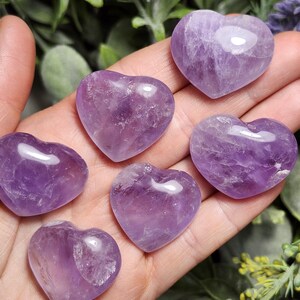 Puede incluir: Seis cristales de amatista en forma de coraz&oacute;n de diferentes tama&ntilde;os y tonos de p&uacute;rpura. Los cristales son lisos y pulidos, con un aspecto natural y nublado.