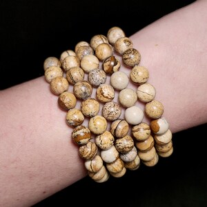 May include: A stack of four beaded bracelets. The beads are round, with a diameter of approximately 0.4 inches. The beads have a mix of tan, brown, and cream colors, with speckled and striped patterns. The bracelets are worn on a wrist.