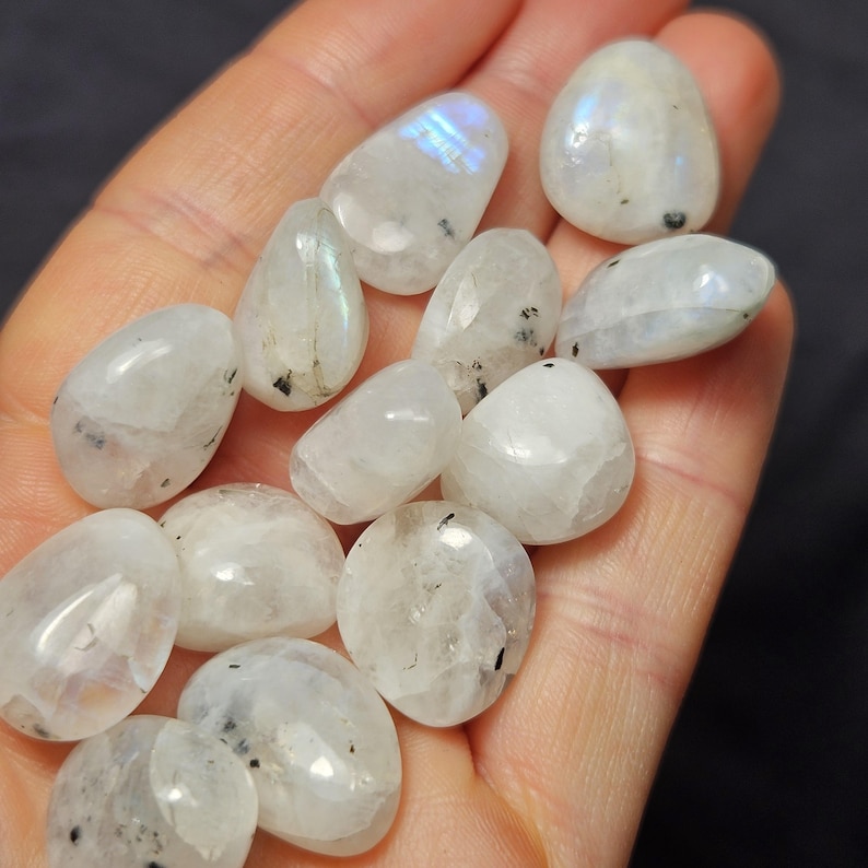 May include: A handful of polished moonstone gemstones in various shapes. The stones are mainly white with subtle iridescent flashes of blue and rainbow colours. Some stones have small black inclusions.