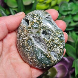 May include: A polished, oval-shaped gemstone with a mix of green, white, and brown hues. The stone has a speckled pattern and several small, open cavities. The gemstone is held in a hand, with a blurred green background.