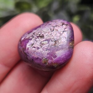 May include: A polished, rounded gemstone with a deep purple hue, accented by shimmering silver and gold inclusions. The stone's surface has a smooth, tactile appearance, with subtle variations in color and texture. The gemstone is held in a hand.