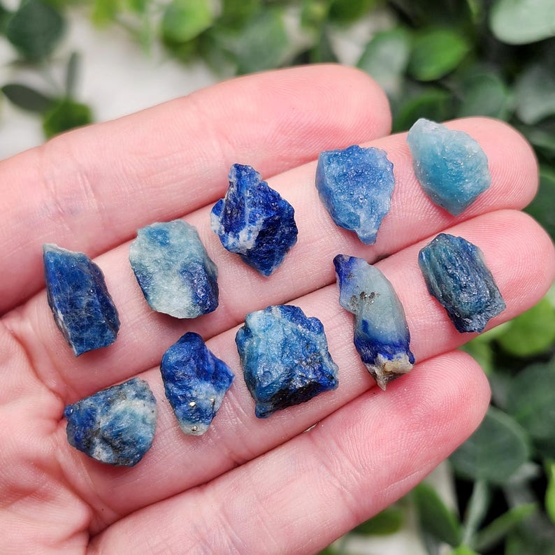 May include: A collection of raw, natural blue crystals in varying shades, from deep indigo to light turquoise. The crystals are irregularly shaped and appear to be held in a hand, showcasing their natural textures and colors. The crystals are a mix of different sizes.