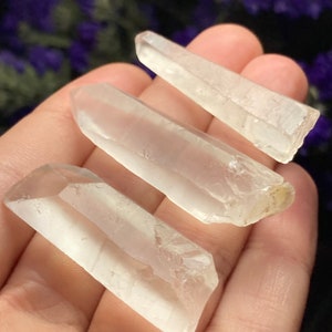 May include: Three clear quartz crystal points of varying sizes, held in a hand. The crystals are smooth and translucent, with a slight shimmer.
