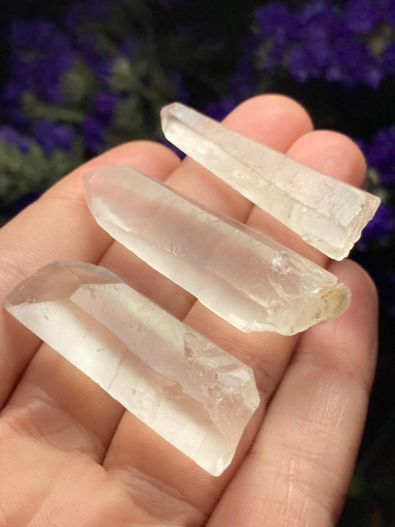 May include: Three clear quartz crystal points of varying sizes, held in a hand. The crystals are smooth and translucent, with a slight shimmer.