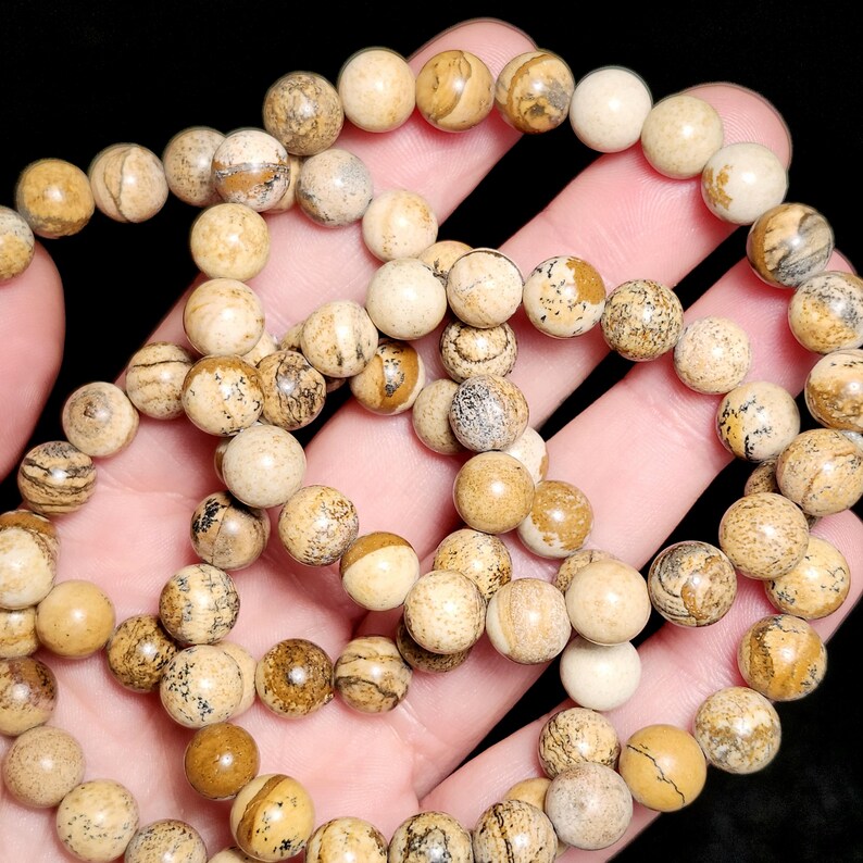 May include: A strand of round, beige and brown jasper beads. The beads have a natural, earthy appearance with varying patterns and shades of tan, brown, and cream. The beads are held in a hand against a dark background.