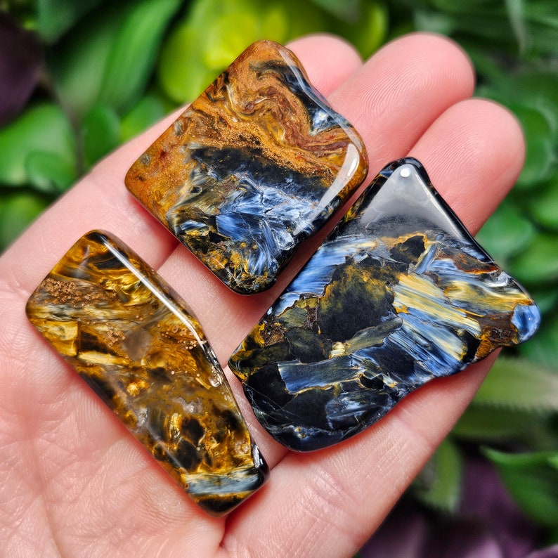 May include: Three blue tiger's eye gemstone cabochons. The cabochons are various shades of blue, gold, and black. The cabochons are smooth and polished.