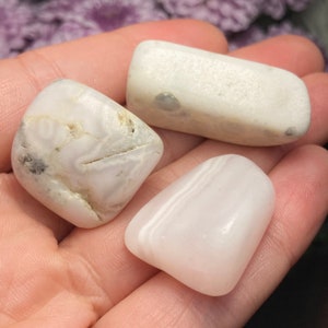 May include: Three polished stones with smooth, rounded surfaces. One stone is opaque white with grey inclusions and linear fissures, another is a rectangular white piece, and the third is a translucent, pale pinkish-white smooth stone.