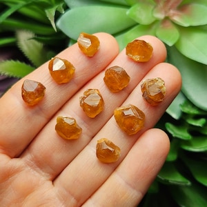 May include: A collection of small, faceted, orange-brown gemstones. The crystals are held in a hand, with a blurred green background. The gemstones have a natural, rough texture and a warm, amber color. The crystals are likely a type of mineral.
