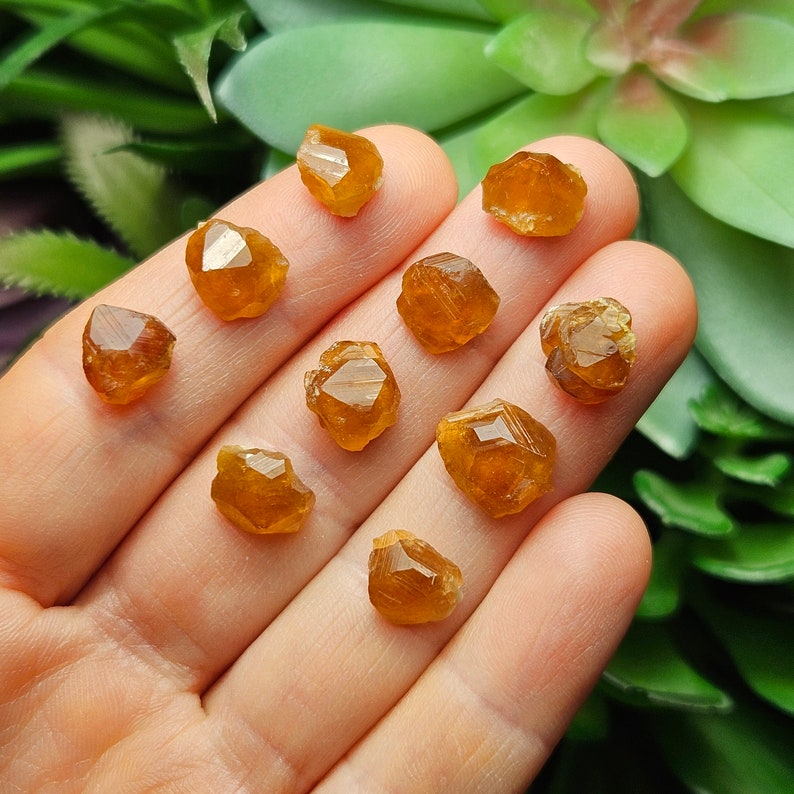 May include: A collection of small, faceted, orange-brown gemstones. The crystals are held in a hand, with a blurred green background. The gemstones have a natural, rough texture and a warm, amber color. The crystals are likely a type of mineral.