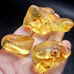 May include: Three polished pieces of translucent golden amber, each featuring irregular shapes and smooth, rounded edges. The stones display internal inclusions, varying shades of yellow and orange, and a glossy, reflective surface texture.