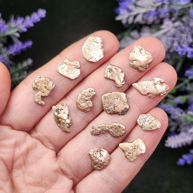 May include: A collection of small, irregularly shaped gold nuggets. The nuggets vary in size and texture, with a shiny, metallic appearance. The nuggets are displayed on a hand, with purple flowers in the background.