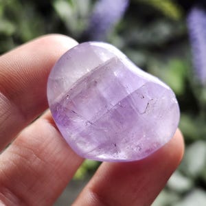 Puede incluir: Un cristal de amatista en forma de coraz&oacute;n con un tono p&uacute;rpura claro y estr&iacute;as naturales. El cristal es liso y pulido, con un borde ligeramente redondeado.