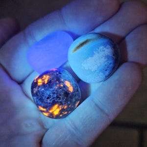 May include: Three polished stones in a hand, illuminated by a blue light. One stone glows with orange and blue patterns, another is light blue with white patches, and the third is a solid blue rectangle. The stones are smooth and rounded.