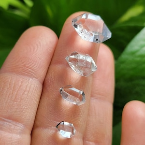 May include: Four clear quartz crystals of varying sizes, arranged in a row on a finger. The crystals are faceted and have a natural, rough appearance.