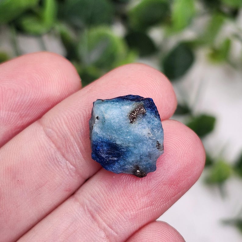 May include: A small, rough-cut gemstone with a mix of blue and light blue hues. The stone has dark blue edges and small flecks of gold. The gemstone is held in a person's hand.