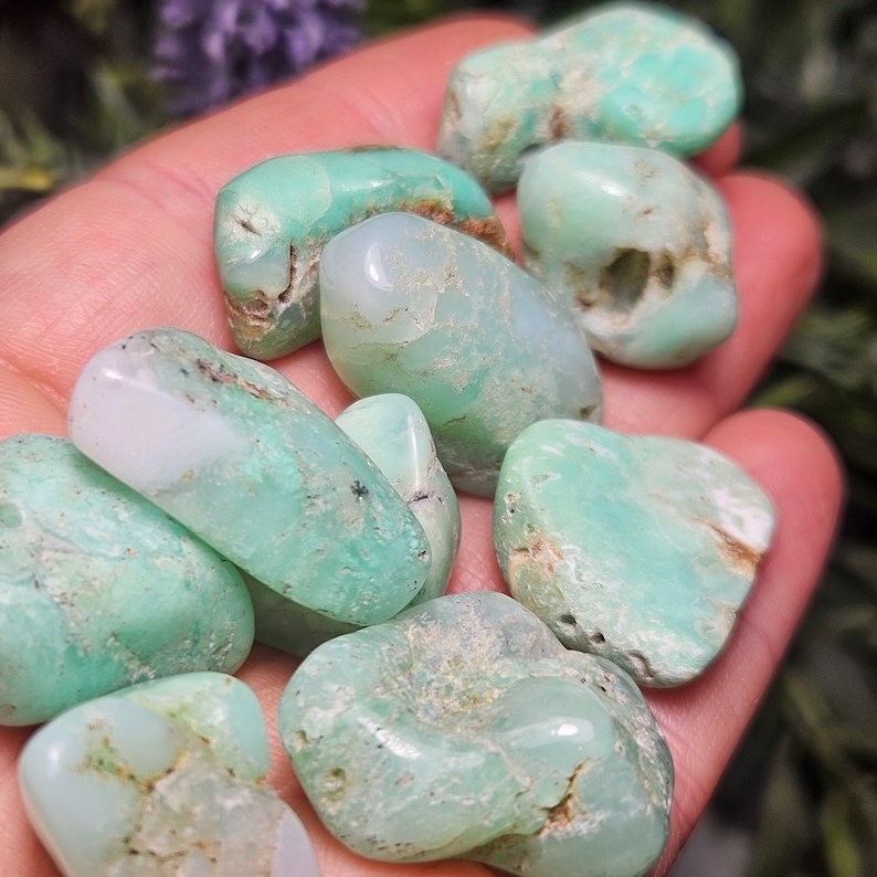 May include: A collection of tumbled chrysoprase stones featuring smooth, polished surfaces in various shades of mint green and pale seafoam. The stones exhibit natural white inclusions, translucent areas, and irregular, rounded shapes.