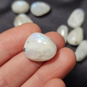 May include: A selection of polished moonstones, oval in shape. The stones are mainly white, with iridescent blue and rainbow flashes. One stone is held in a hand, with others in the background.