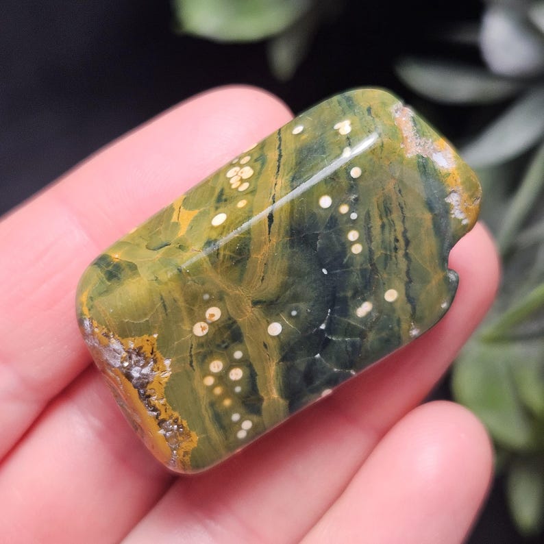 May include: A polished, rectangular gemstone with a unique pattern of green, yellow, and white. The stone has a natural, organic appearance with a smooth, glossy finish. The gemstone is held in a hand, showcasing its size and shape.