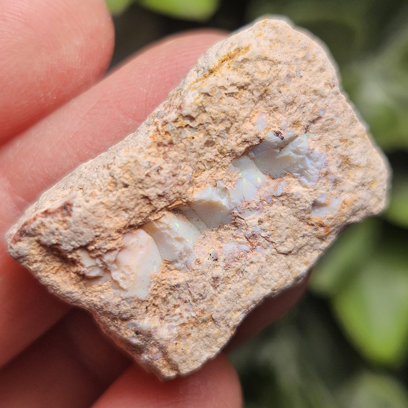 May include: A raw, unpolished rock specimen featuring a central vein of iridescent white opal embedded within a rough, tan-colored matrix. The stone displays a textured, earthy exterior with visible mineral deposits and subtle flashes of color.