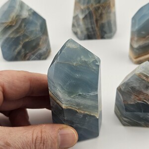 May include: Several blue calcite crystal towers with natural patterns are displayed. The crystals have a layered appearance with shades of blue, brown, and white. One crystal is held by a hand, showcasing the polished facets and unique colour variations.