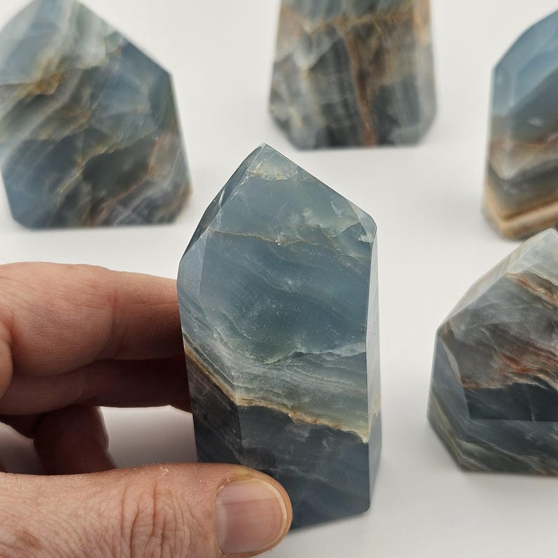 May include: Several blue calcite crystal towers with natural patterns are displayed. The crystals have a layered appearance with shades of blue, brown, and white. One crystal is held by a hand, showcasing the polished facets and unique colour variations.