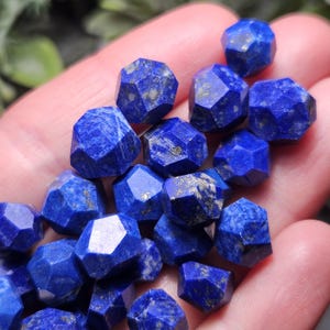 May include: A collection of small, faceted lapis lazuli gemstones. Each stone features a deep blue color with natural golden pyrite inclusions and a multi-sided geometric cut, creating a reflective surface across the various individual pieces.